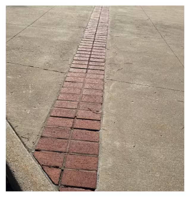 Concrete walkway with a narrow strip of red brick pavers inset down the center, showing visible wear and uneven joints.