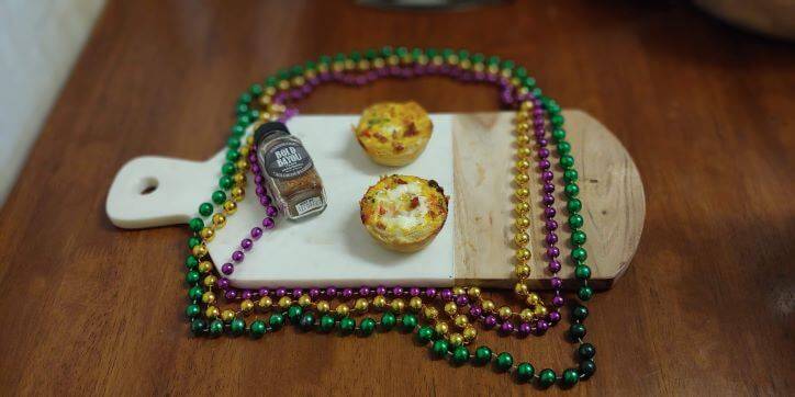 A cutting board with FreshJax Organic Bold Bayou Cajun Spice next to cajun mini quiches. Mardi Gras beads on table. 
