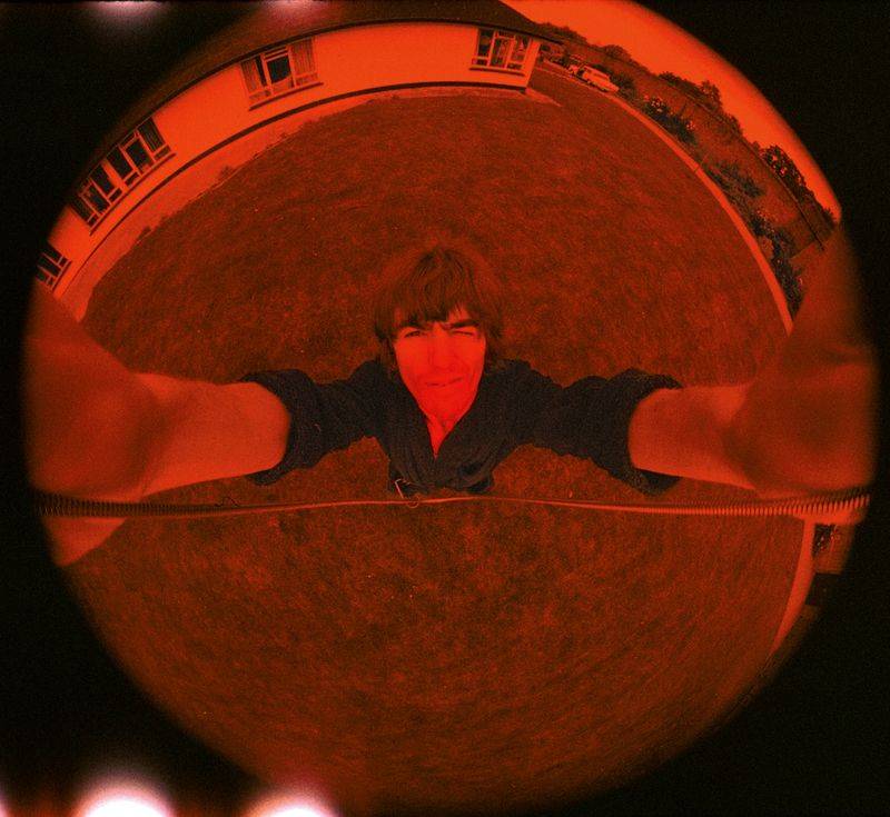 A red and black fish-eye style photograph of a young George Harrison, seen from above. Harrison is taking the photo with a camera that he's holding above himself. His face is in the center of the circular image. His arms are stretched out on either side t