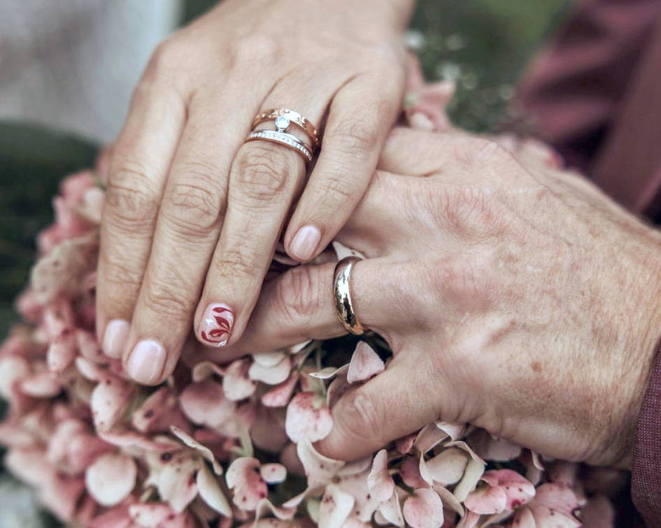 La main gauche d'une femme parée de bagues de fiançailles et de mariage posée sur celle d'un homme parée d'un jonc en or au-dessus d'un bouquet de mariage.