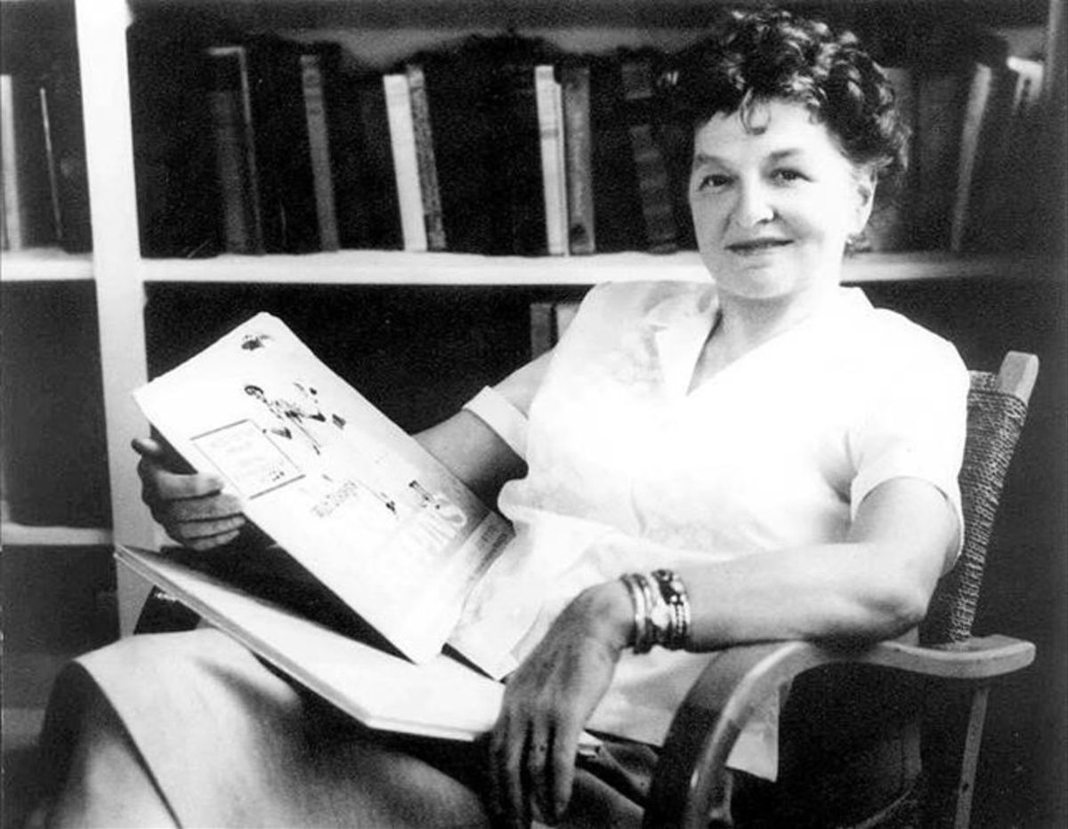 Black and white image of Travers sitting on a wodden chair in front of a bookshelf, smiling calmly with a book on her lap.