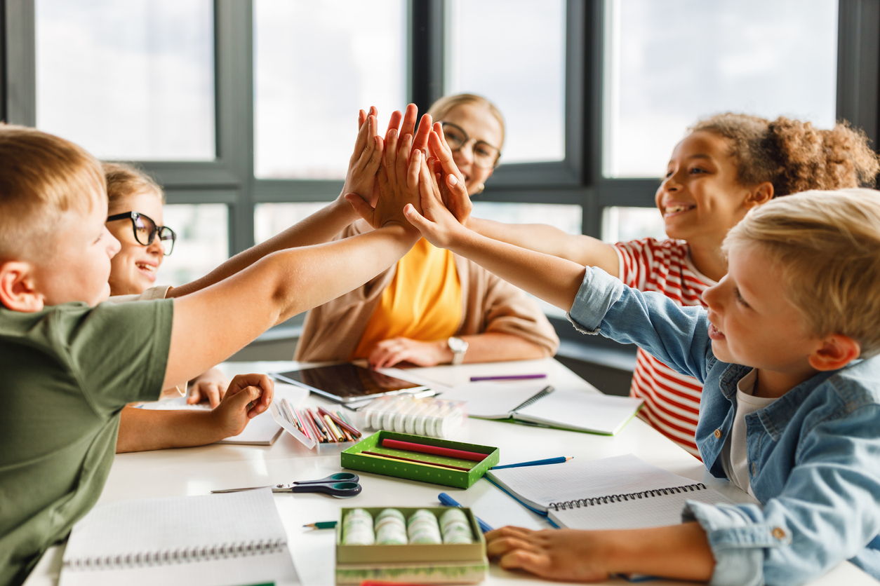 Four children sitting around a table with school supplies, smiling and giving a group high five, with an adult woman in glasses watching and smiling in the background. Large windows let in natural light.