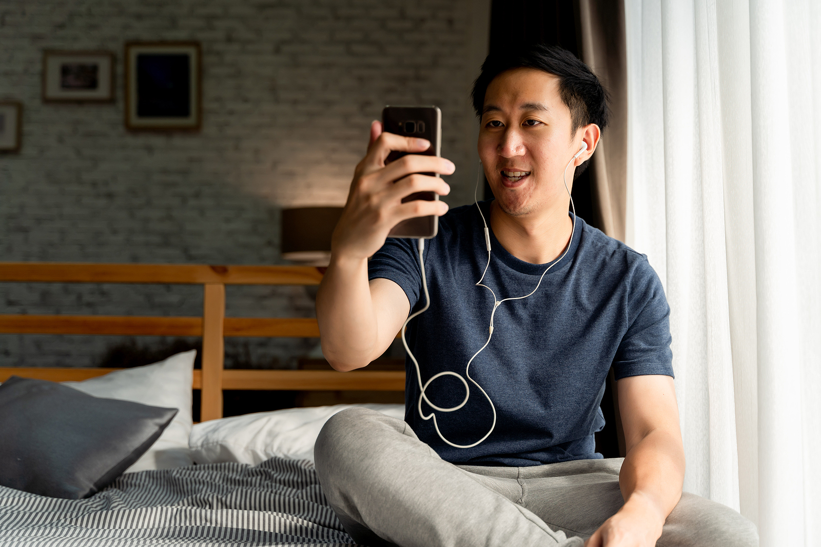 Attractive young Asian man sitting on his bed and smiling while video chatting with someone on his phone.