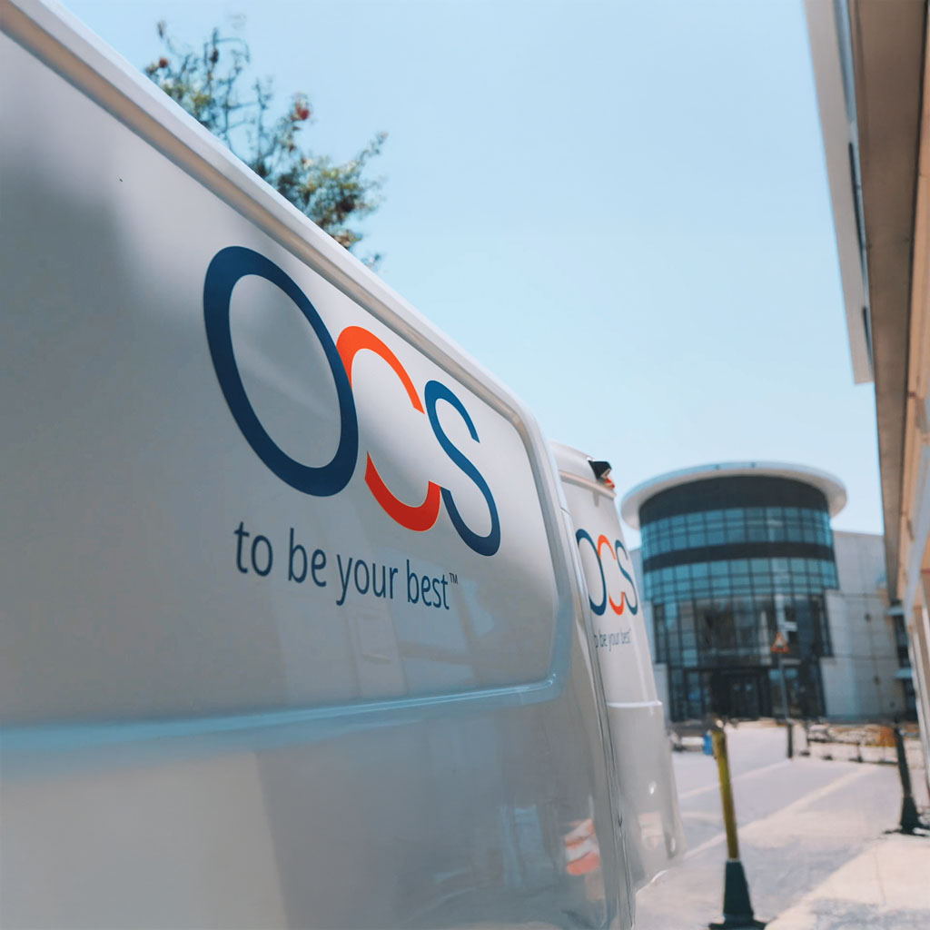A close-up of a white vehicle with the OCS to be your best logo, parked on a sunny street near a modern, glass-fronted building.