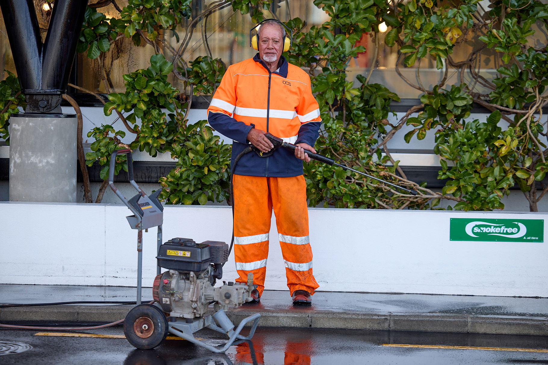 A worker in bright orange safety gear stands on a wet pavement holding a power washer, with green bushes and a smoke free sign in the background. A power washer machine is positioned in front of him.