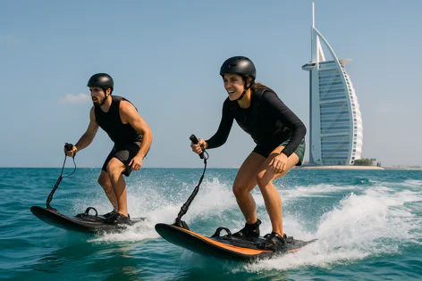 Two smiling people riding electric surfboards on a sunny day in the ocean, with the iconic Burj Al Arab building visible in the distance.