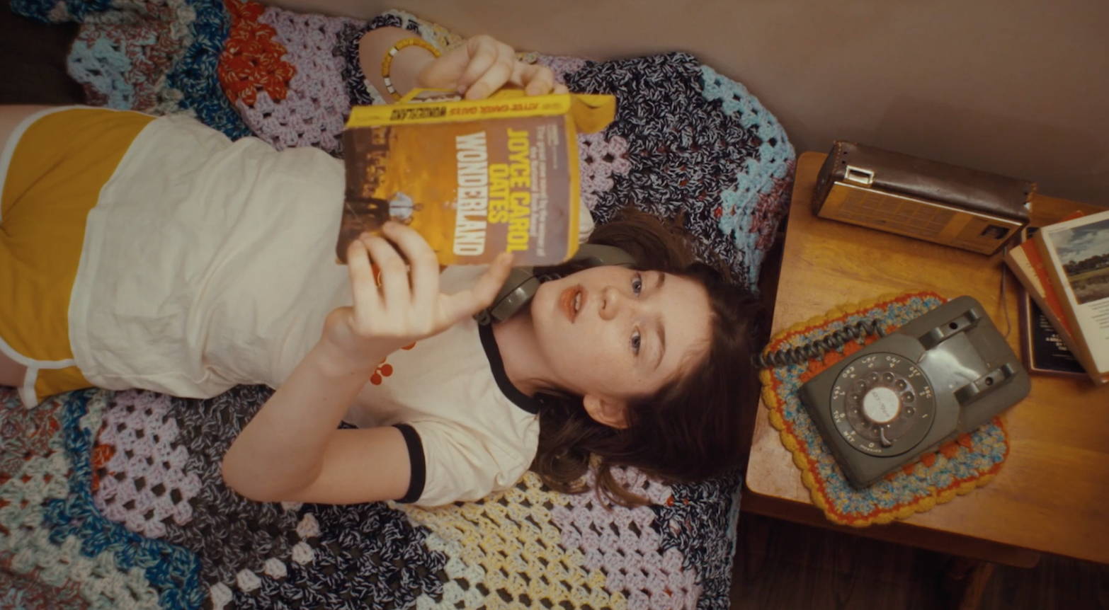 Girl laying on her bed, talking on a 1970s phone, holding a book
