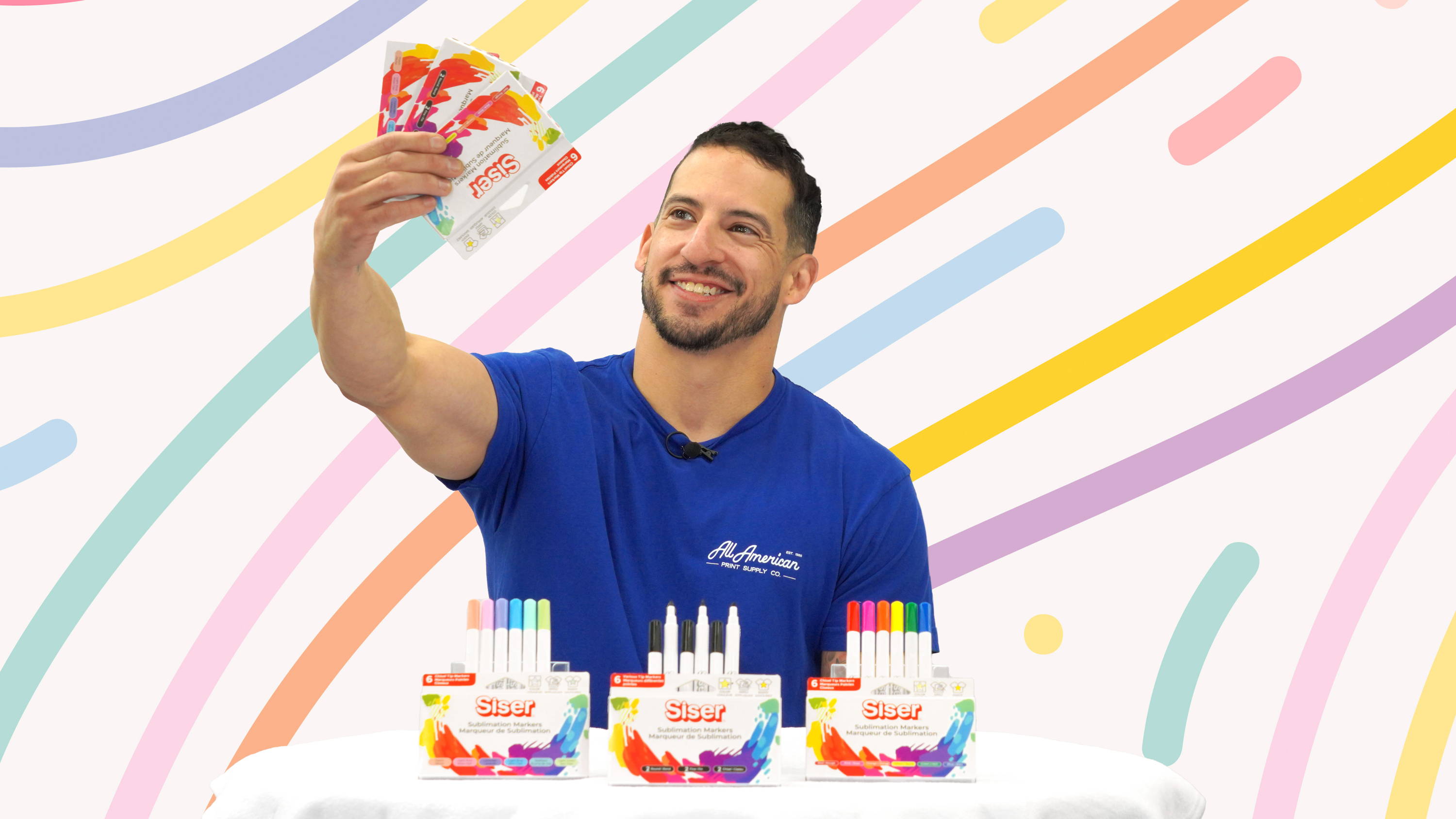 All American Print Supply Co's Estevan Romero holds up a pack of the all new Siser Sublimation Markers. Opened packs in front of him display the varying tip shapes, the primary color pack, and the pastel color pack. Graphic color strokes appear behind Estevan.