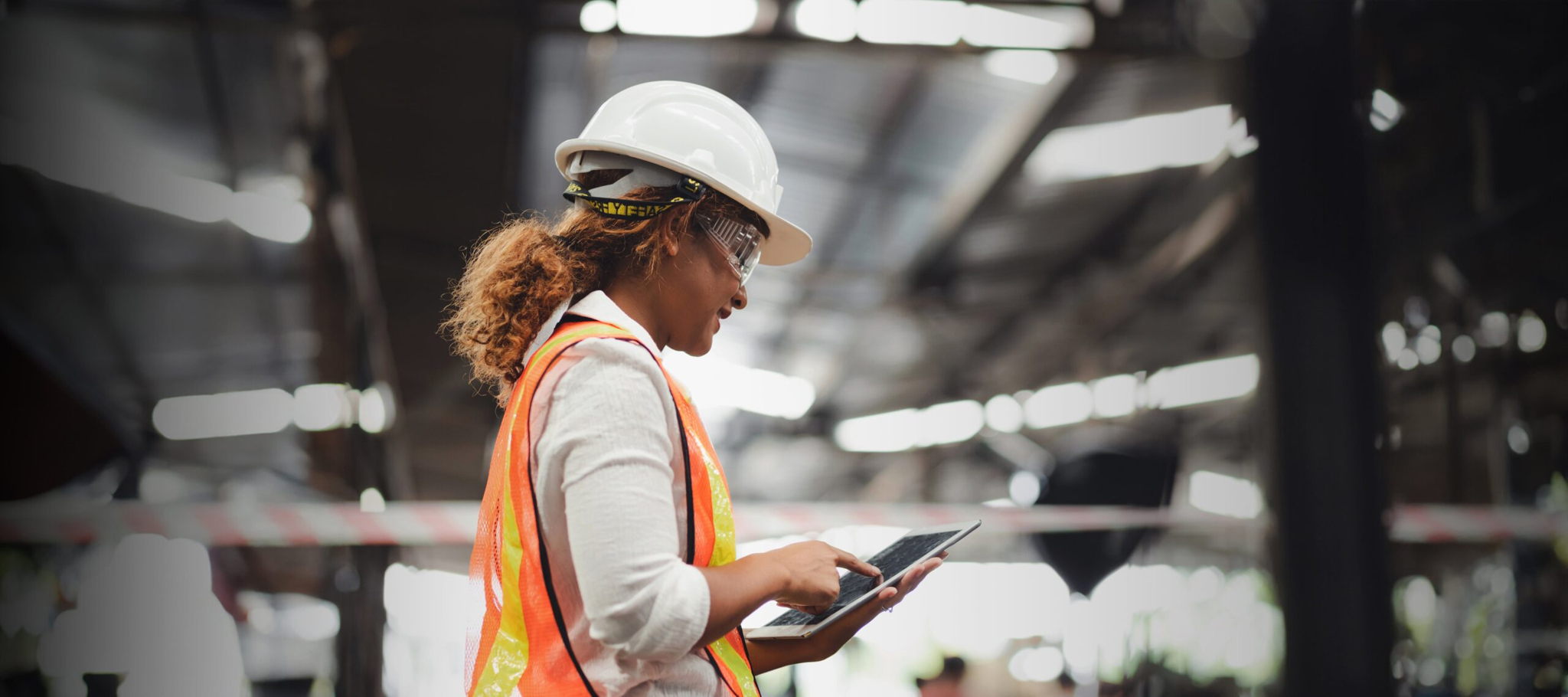A woman in an orange safety vest and white hard hat uses a tablet in an industrial setting. She is also wearing safety goggles. The background is a blurred view of a factory or warehouse with metal structures.