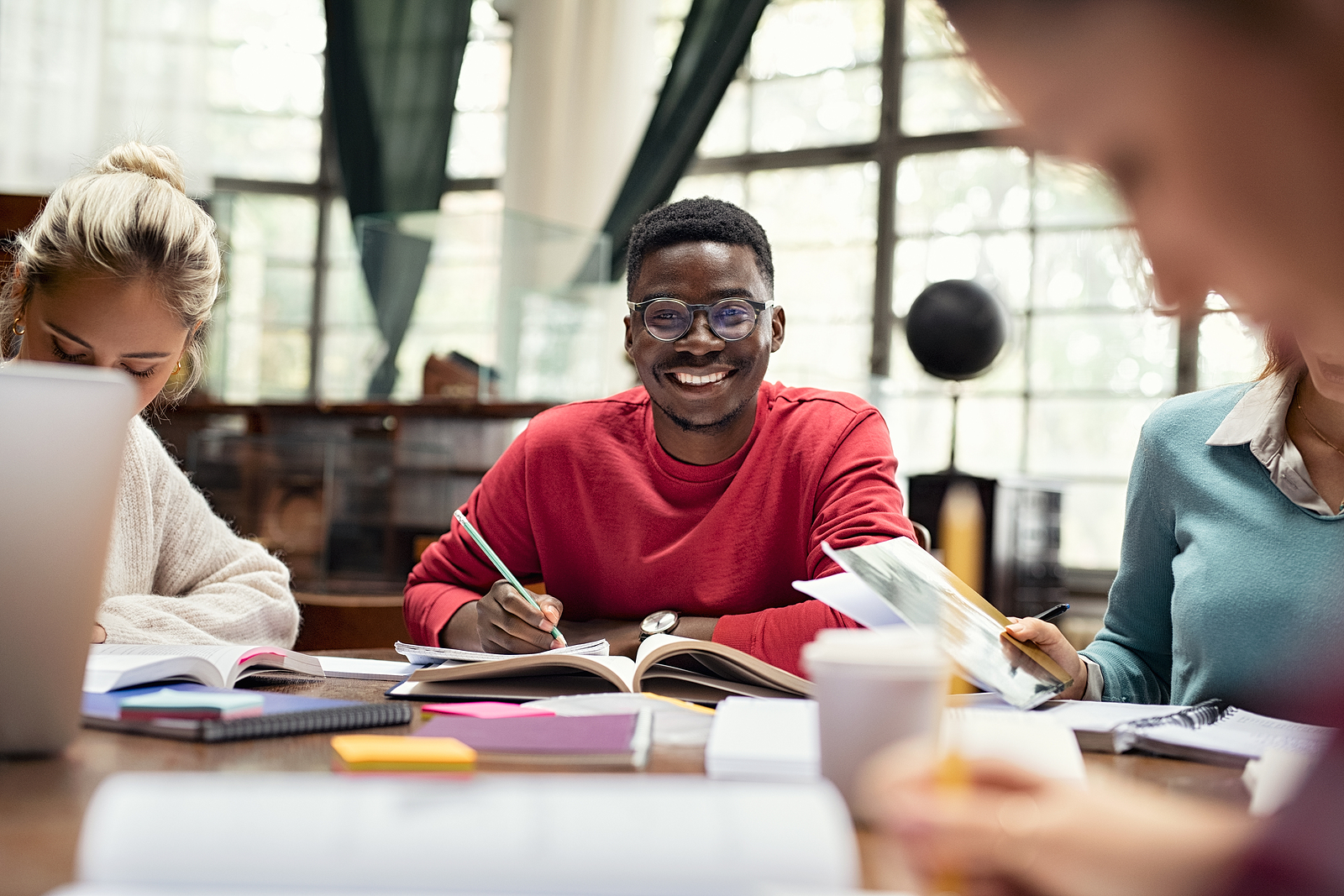 Image of a young black man smiling and writing on a journal next to other classmates doing the same.