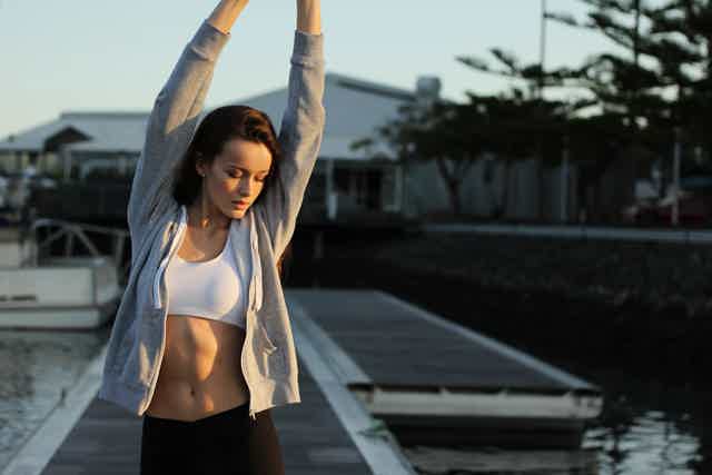 Athletic woman stretching arms overhead on a dock by water, wearing white sports bra and gray zip hoodie.
