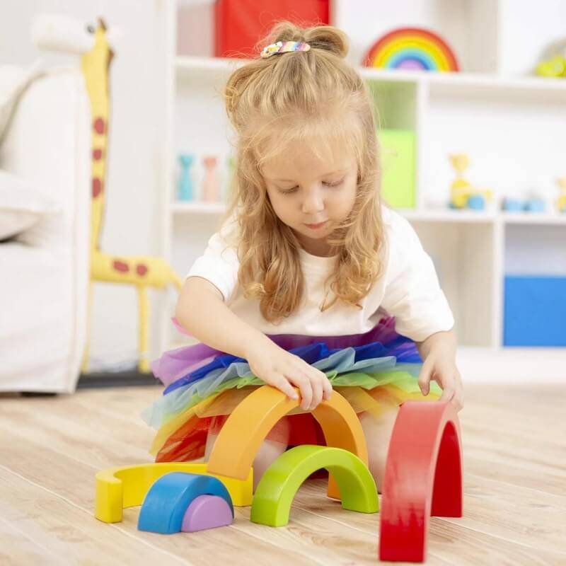 Une jeune fille aux cheveux blonds, vêtue d'une jupe tutu colorée, joue avec un arc-en-ciel Montessori d'InvenToy dans une salle de jeux joliment décorée.