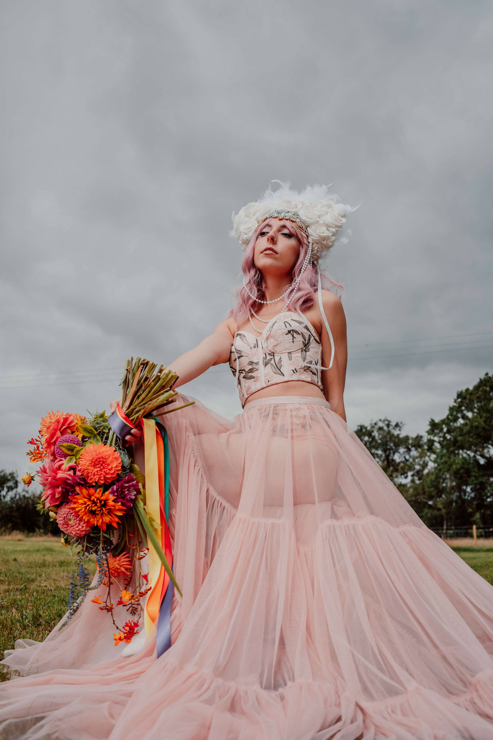 Image of a bride at a festival. She is wearing an embroidered modern bridal outfit, white head piece instead of a veil, and has biodegradable blue, pink, and iridescent glitter on her face. She's holding a bright and colourful bridal floral bouquet.