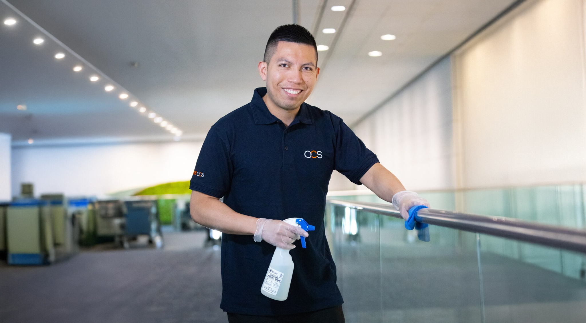 A man wearing a blue polo shirt and gloves is smiling while cleaning a glass railing with a spray bottle. He stands in a large, well-lit indoor area with a modern design and soft overhead lights.