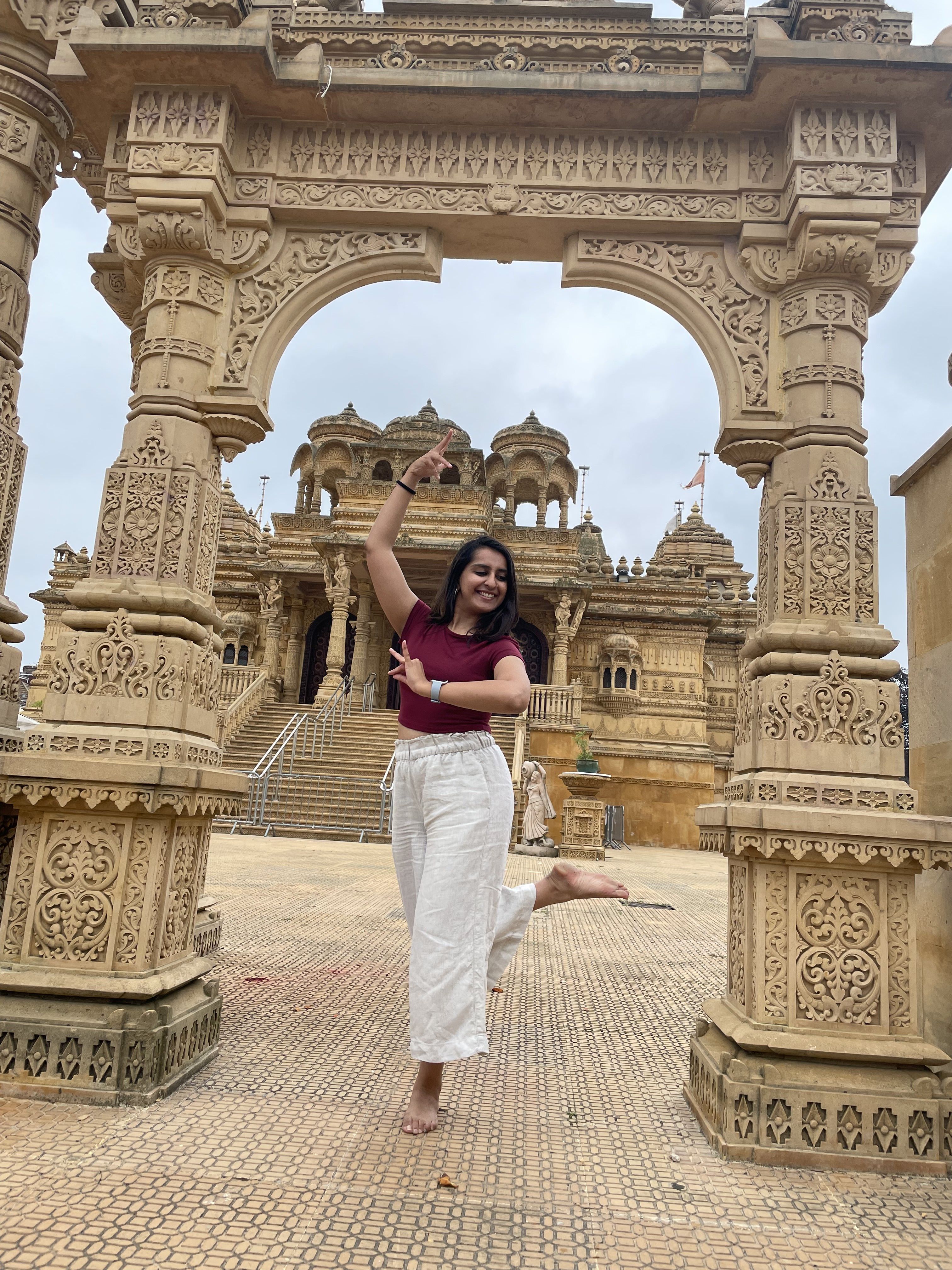 Yashodhra is wearing white trousers and a burgundy t-shirt. She strikes a traditional Indian dance pose in front of the Hindu Temple in Wembley, London.