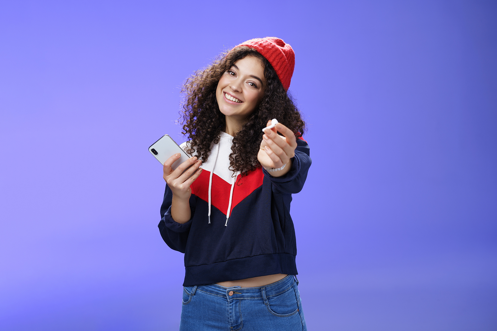 Portrait of friendly charming and cute woman in warm beanie extending arm towards camera to share an earphone, holding a smartphone.