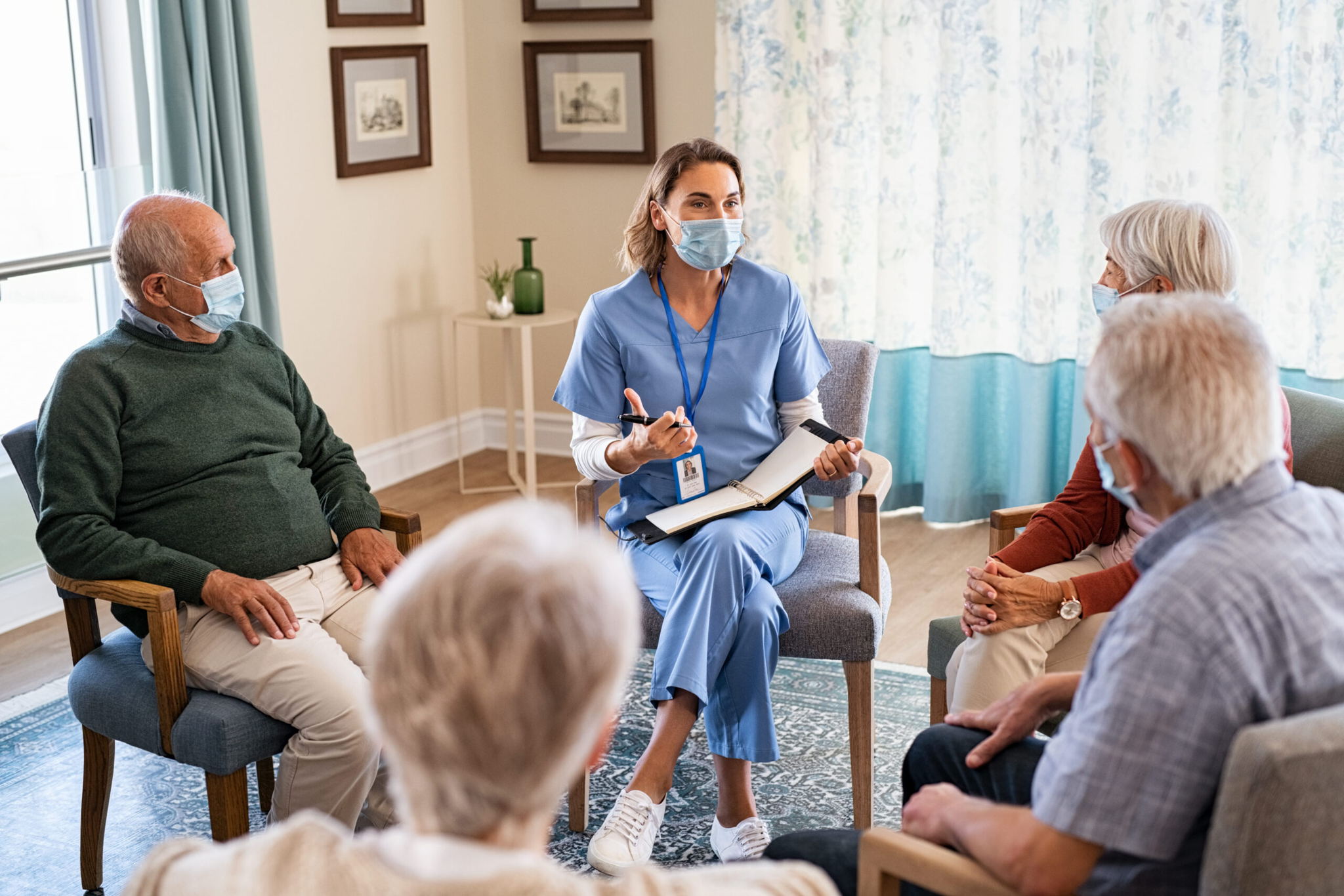 A nurse wearing scrubs and a face mask leads a discussion with a small group of older adults seated in a circle in a well-lit room, all wearing masks and engaged in conversation.