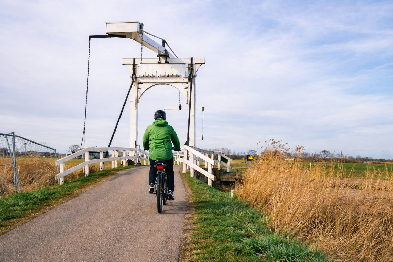 E-biker in groene jas rijdt van achteren gezien over een smal asfaltpad het witte houten ophaalbruggetje op in het Nederlandse polderlandschap.