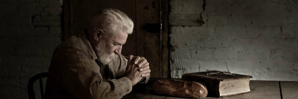 An older man praying at his table next to his bible.