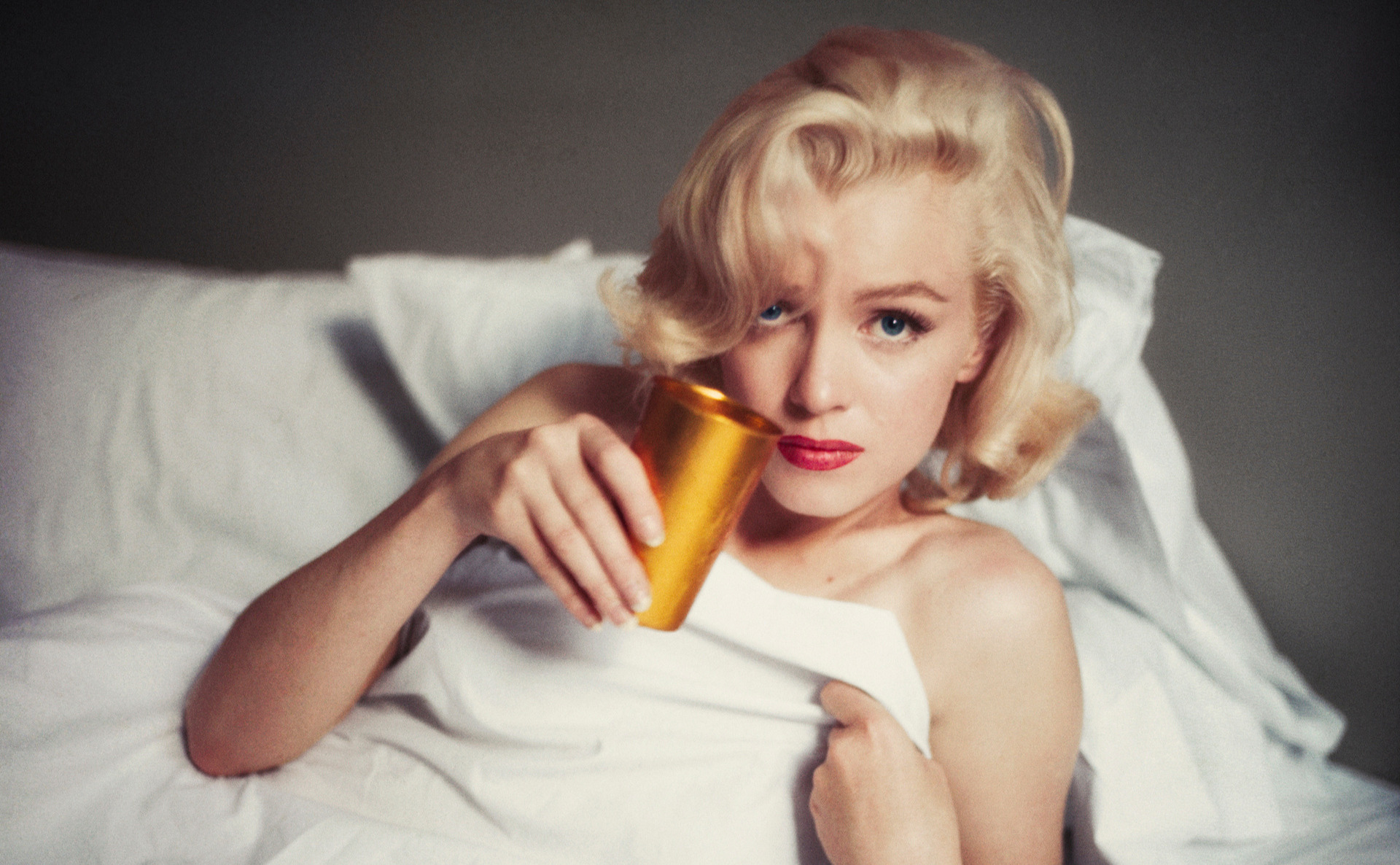 Marilyn Monroe laying a white bed, covered only with blanket, holding a golden cup.