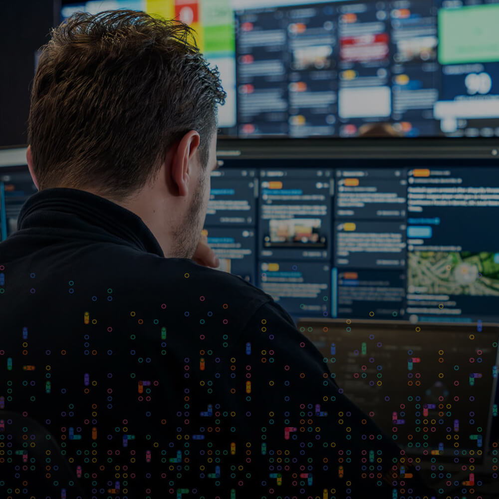 A person with short brown hair sits at a desk, looking at multiple computer monitors displaying various dashboards and information screens in a dark, high-tech setting.