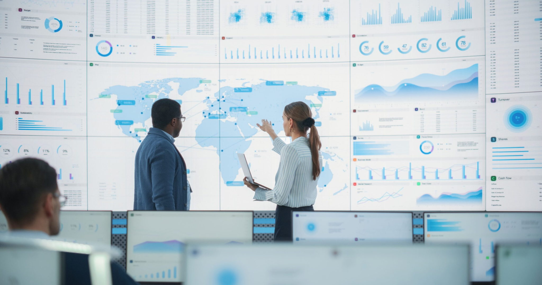 Two professionals discuss data displayed on large screens showing world maps and graphs in a modern office or control room, while other people work at computer stations in the foreground.