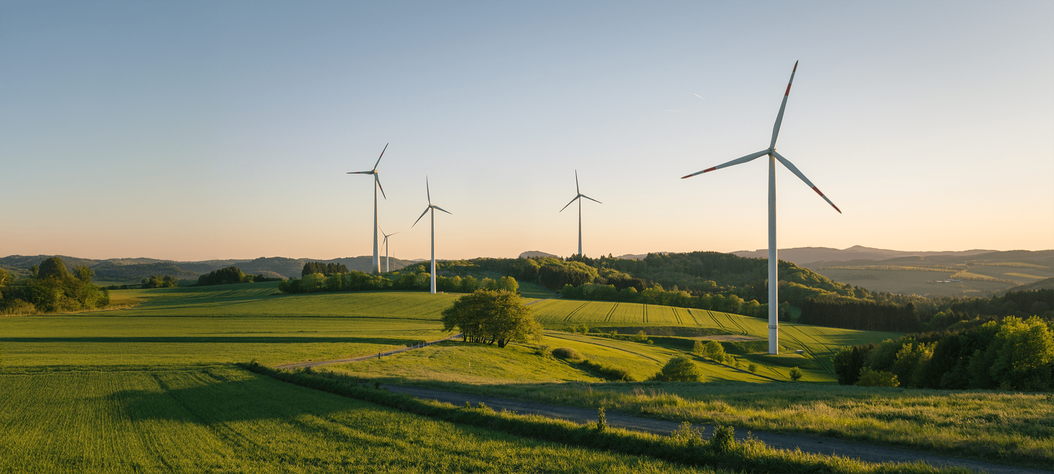 Four large wind turbines stand on green rolling hills under a clear sky at sunset, surrounded by fields, trees, and distant hills, showcasing a peaceful rural landscape with renewable energy.