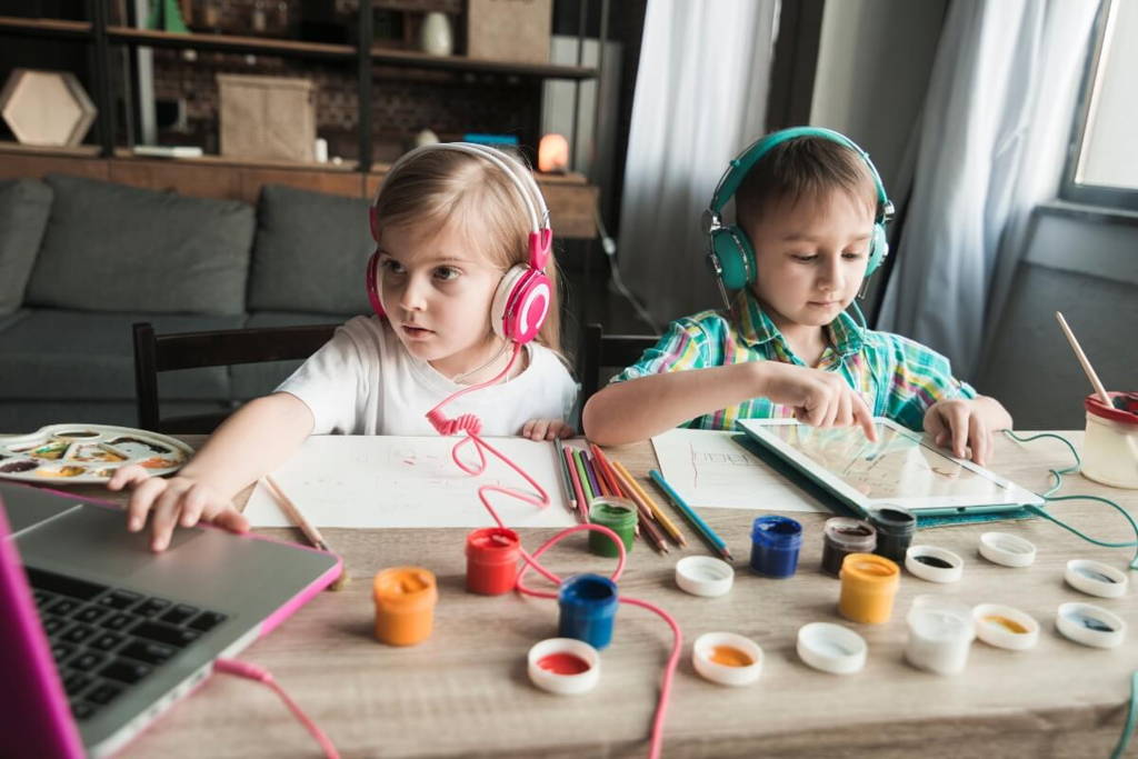 Deux jeunes enfants portant des écouteurs colorés participent à un cours d'art en ligne, utilisant une tablette et un portable tout en étant entourés de peintures, crayons et fournitures artistiques.