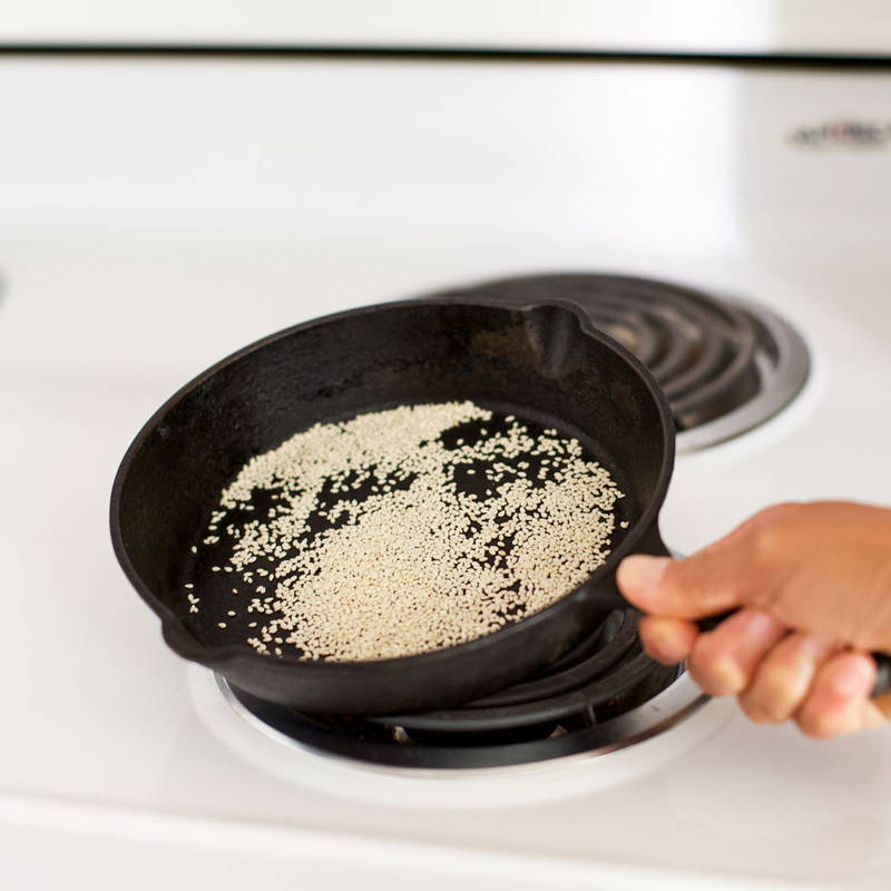 A cast iron skillet over a white stove top is filled with sesame seeds which are being toasted