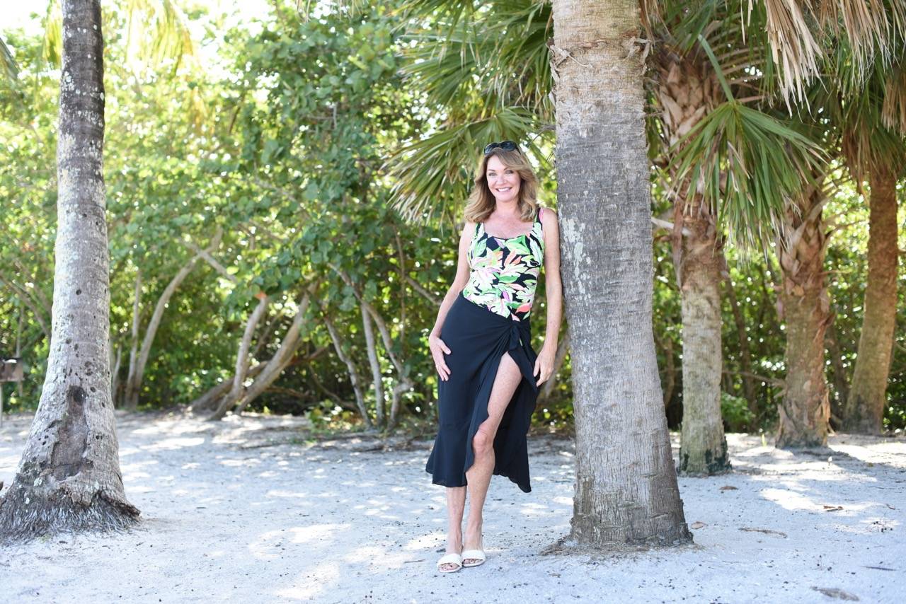 A woman in a rainforest rainbow swimsuit with a black sarong lounges next to a palm on the beach.