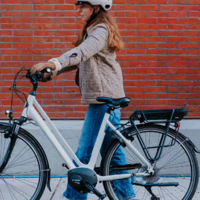 a woman walking next to her electric city bike in front of a red brick building