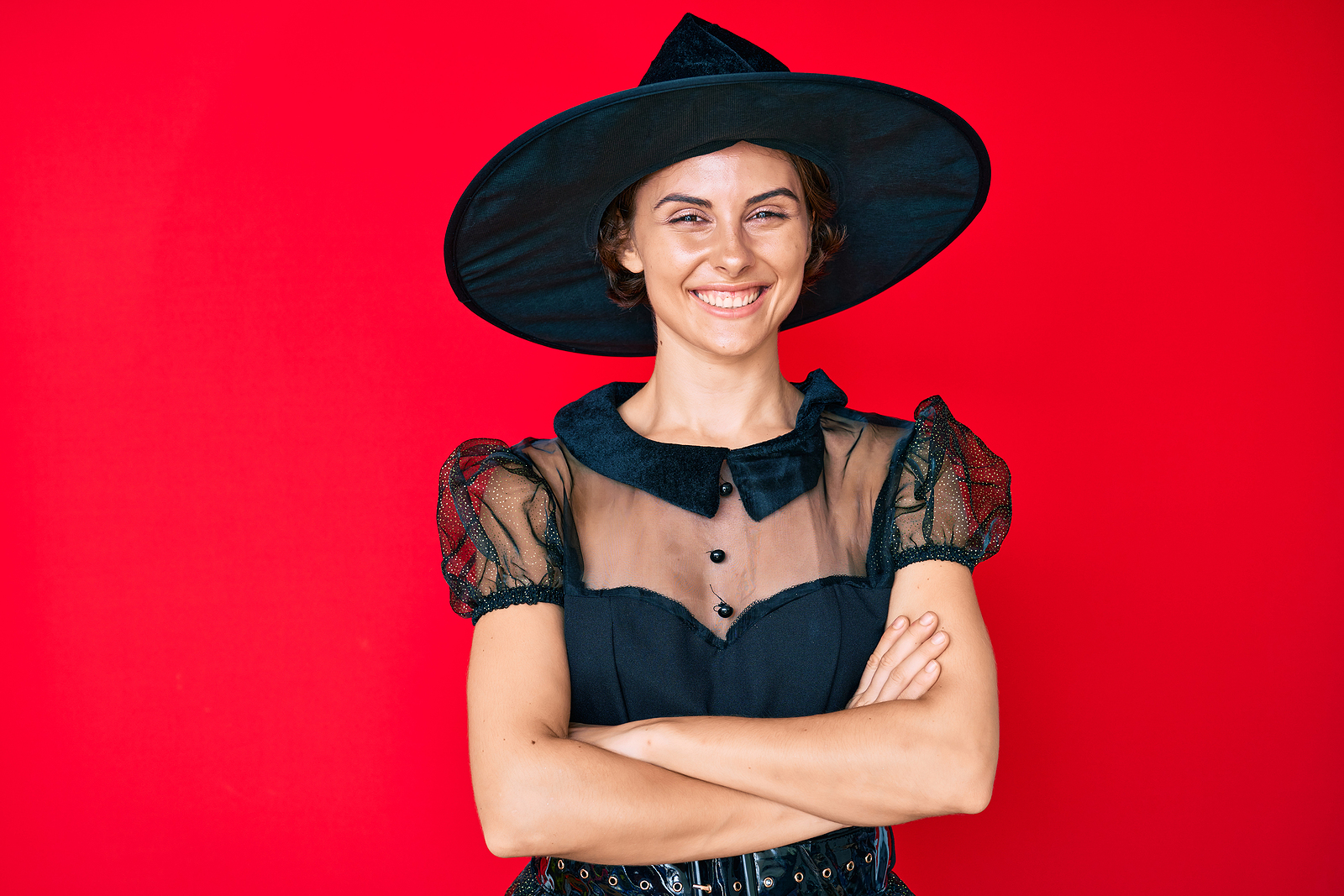 A confident woman with a witch outfit and hat crosses her arms and smiles.
