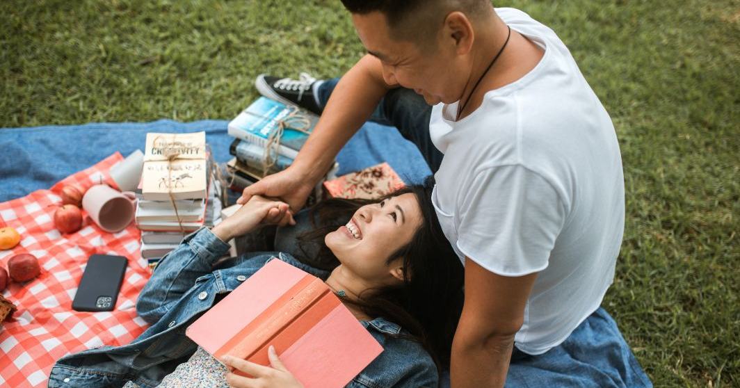 An Asian couple lay on the grass having a picnic together and laughing.