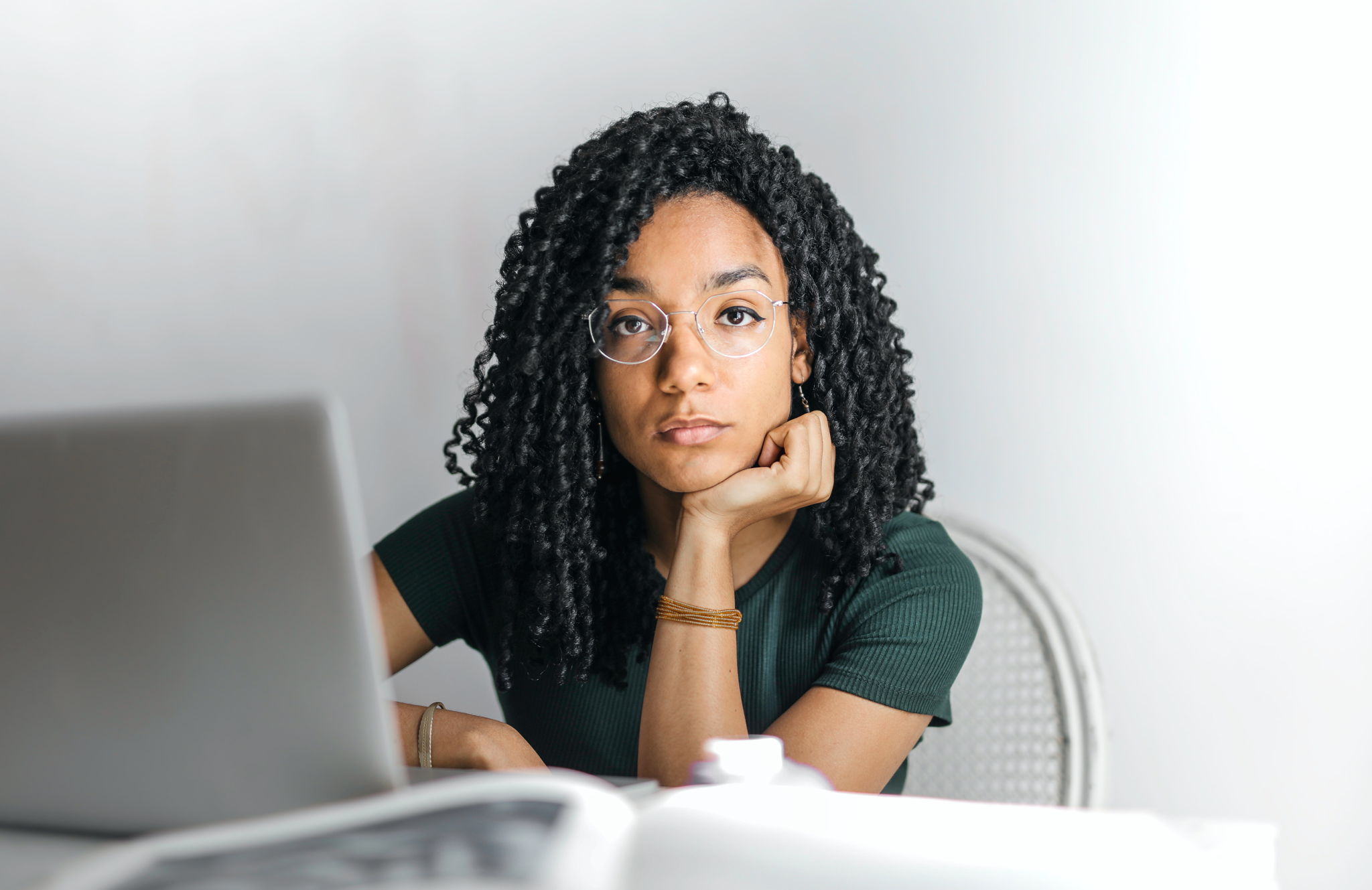 An afro-american girl with curly hair sit in front of a laptop with a serious thinking expression.