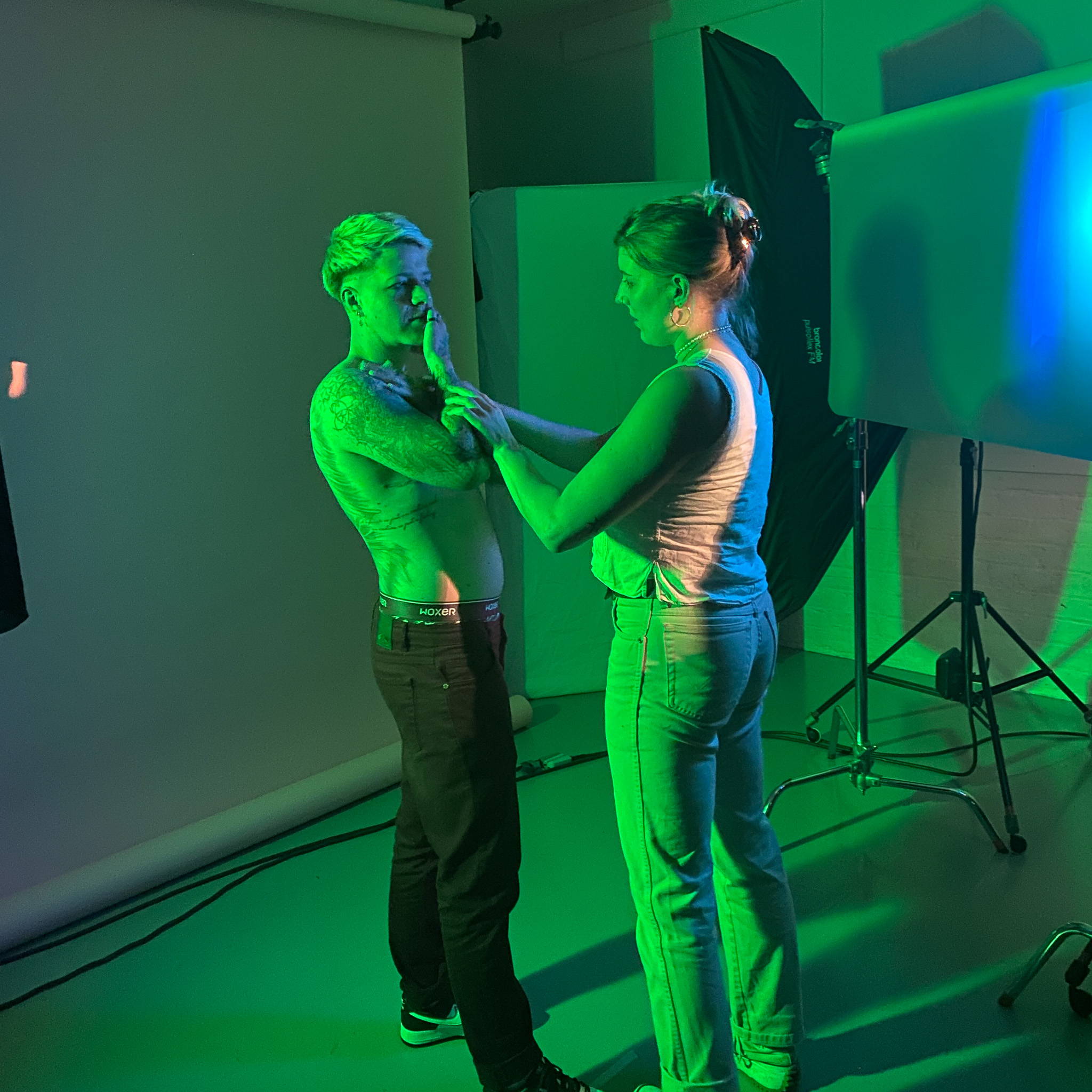 Model TJ stands topless to the left of the image facing stylist Jessie who is holding their arm in pose. They are both bathed in green and blue lighting and in the background we can see a large white photography backdrop, lighting equipment and tripods. 