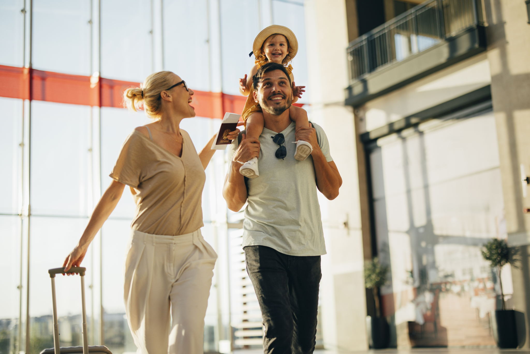 A smiling family walks through a bright airport terminal. An adult carries a suitcase, another adult carries a child on their shoulders. The child is wearing a hat and smiling happily. Sunlight streams in through large windows.