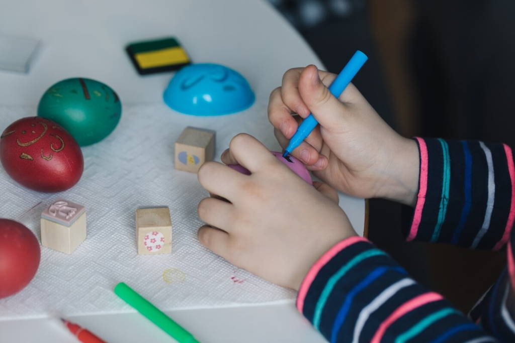 Un enfant décorant un œuf de Pâques en plastique avec un marqueur bleu à une table, entouré d'œufs colorés, de tampons et de marqueurs.