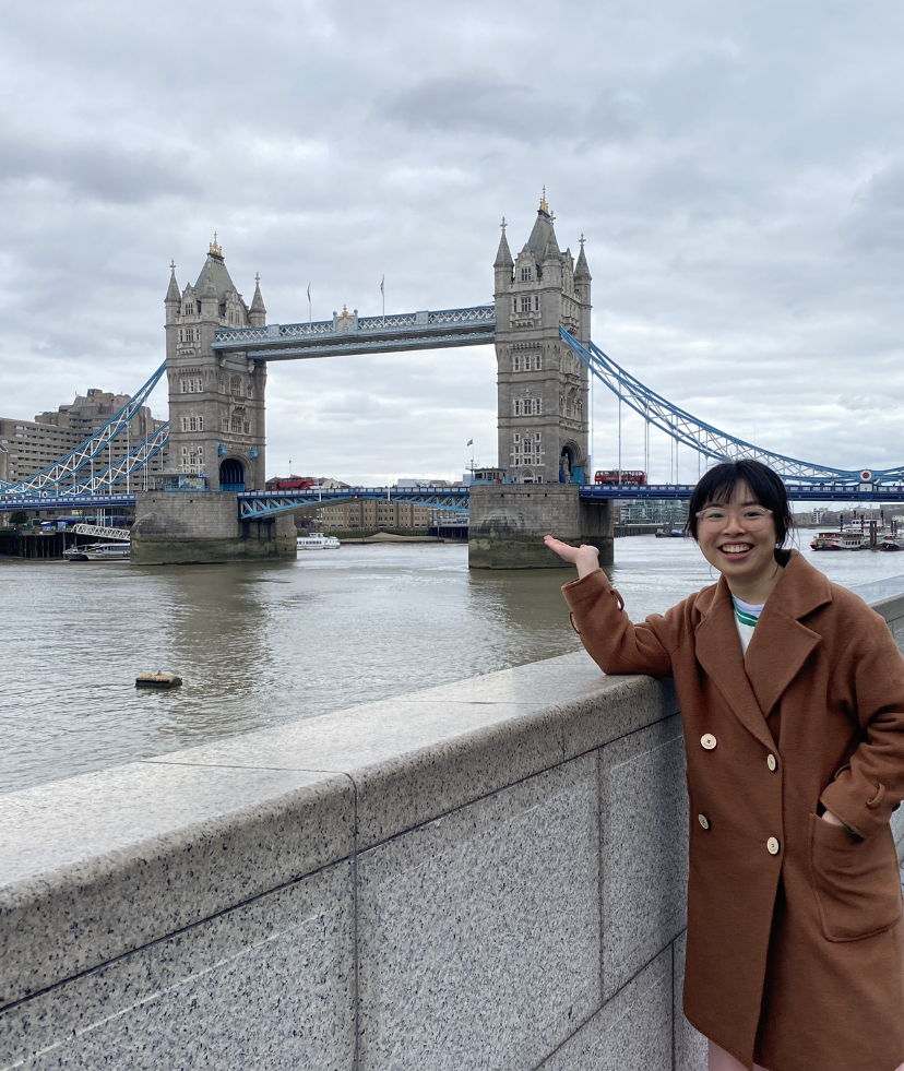 Lan stands in front of Tower Bridge in London wearing a winter coat and smiling, while pointing us towards this London landmark.