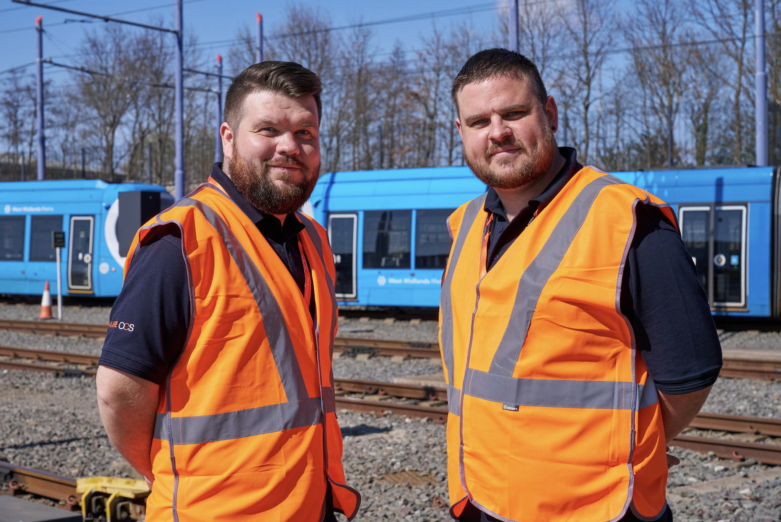 Two men working for OCS rail cleaning wearing bright orange safety vests stand on a railway track with blue trains and trees in the background on a sunny day.