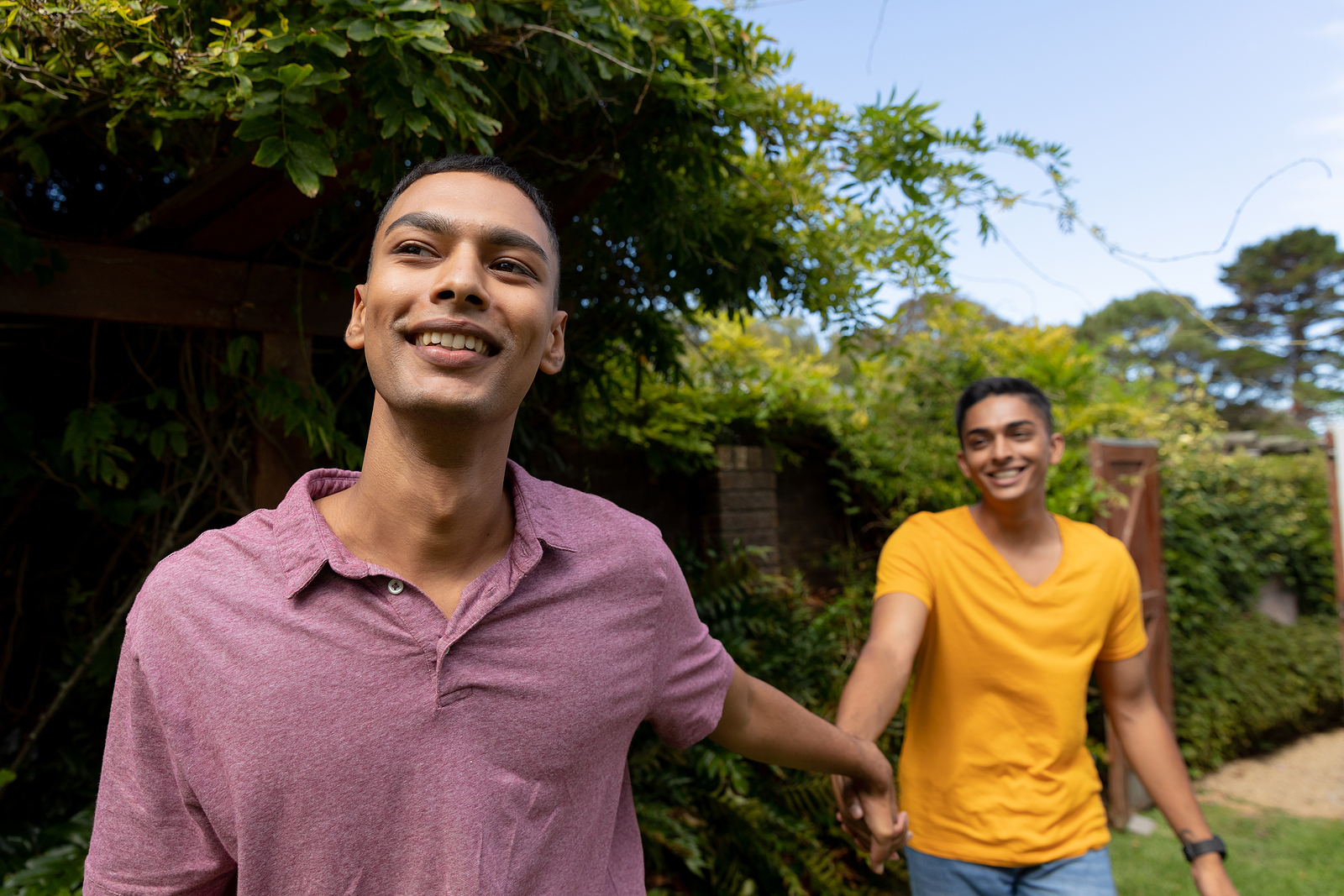 A multi ethnic couple of guys laughing and walking holding by the hand with trees behind them.