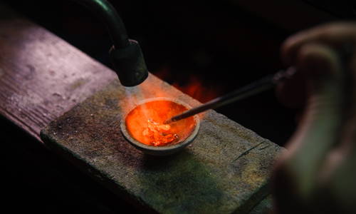 The fire of a torch makes metal liquid in a jeweler's crucible.