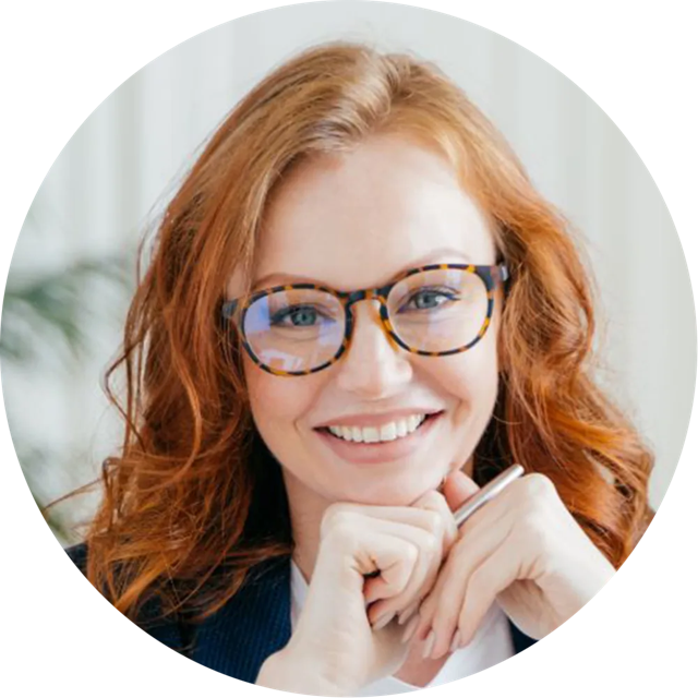Smiling red-haired woman with tortoiseshell glasses, resting chin on hands.