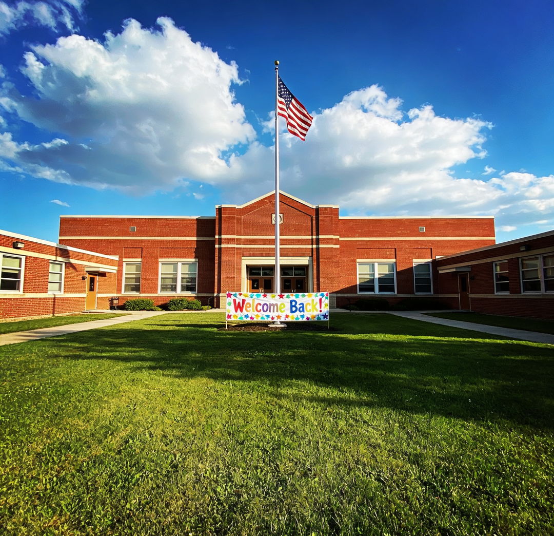 Wide exterior photograph of a well-maintained red brick elementary school building with an American flag flying out front, green lawn, and a colorful welcome banner near the entrance. Bright blue sky with fluffy white clouds, late morning sunlight, 35mm lens, establishing shot conveying stability and community pride.