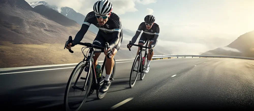 Zwei Radfahrer in Rennradkleidung fahren schnell auf einer bergigen Landstraße. Im Hintergrund sind schneebedeckte Berge und ein klarer Himmel zu sehen, die eine malerische Kulisse für ihre sportliche Fahrt bieten.