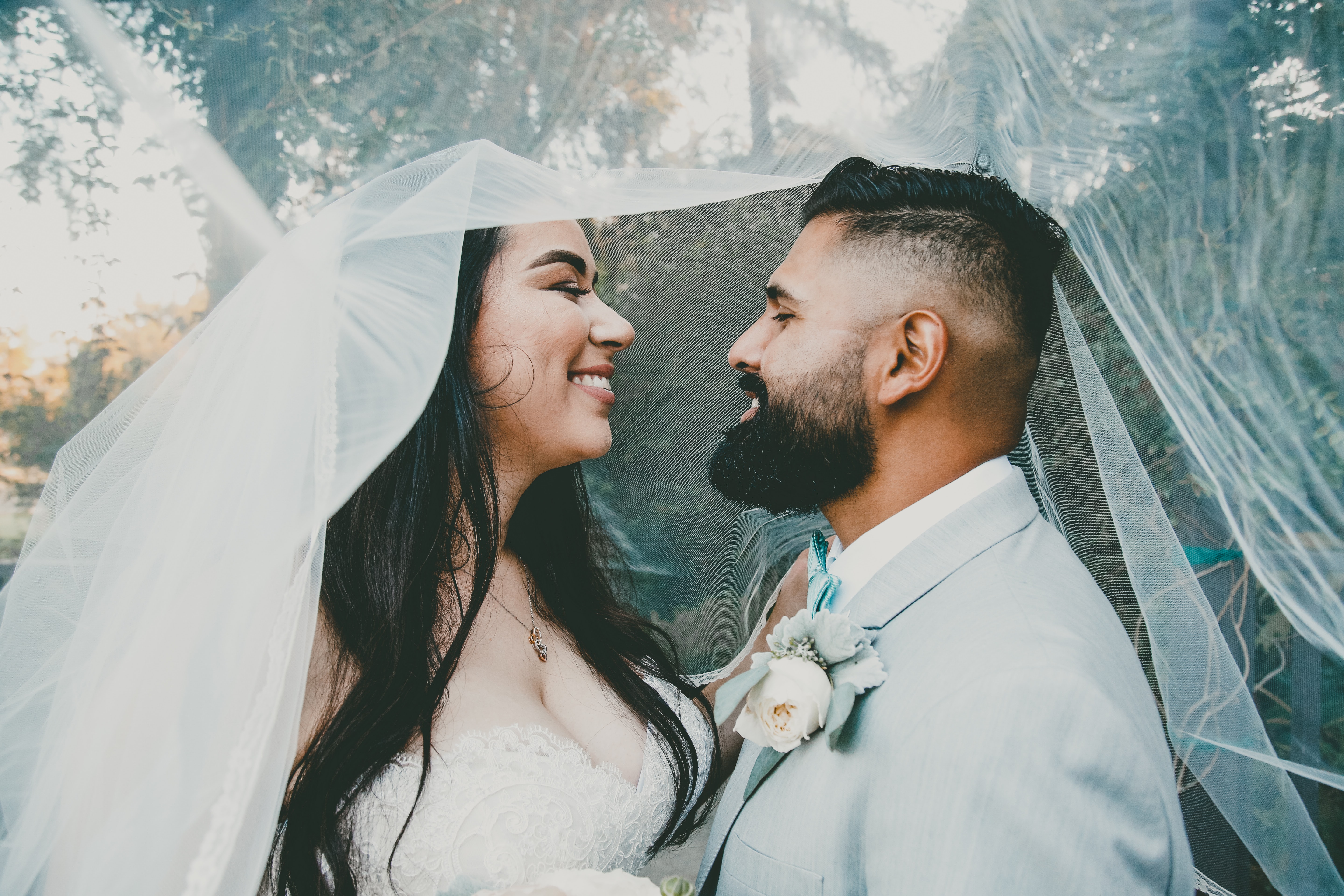 A couple wearing wedding outfits covered in the veil of the bride and smiling at each other.