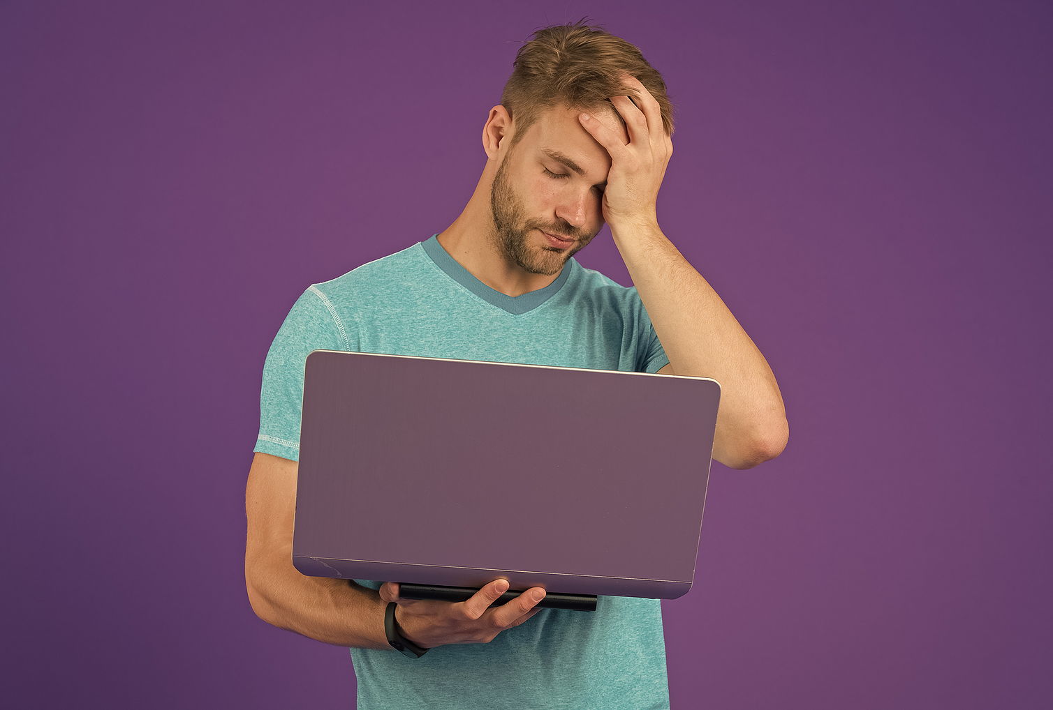 A tired man holds his computer in one hand and his head in the other worried against a purple background.