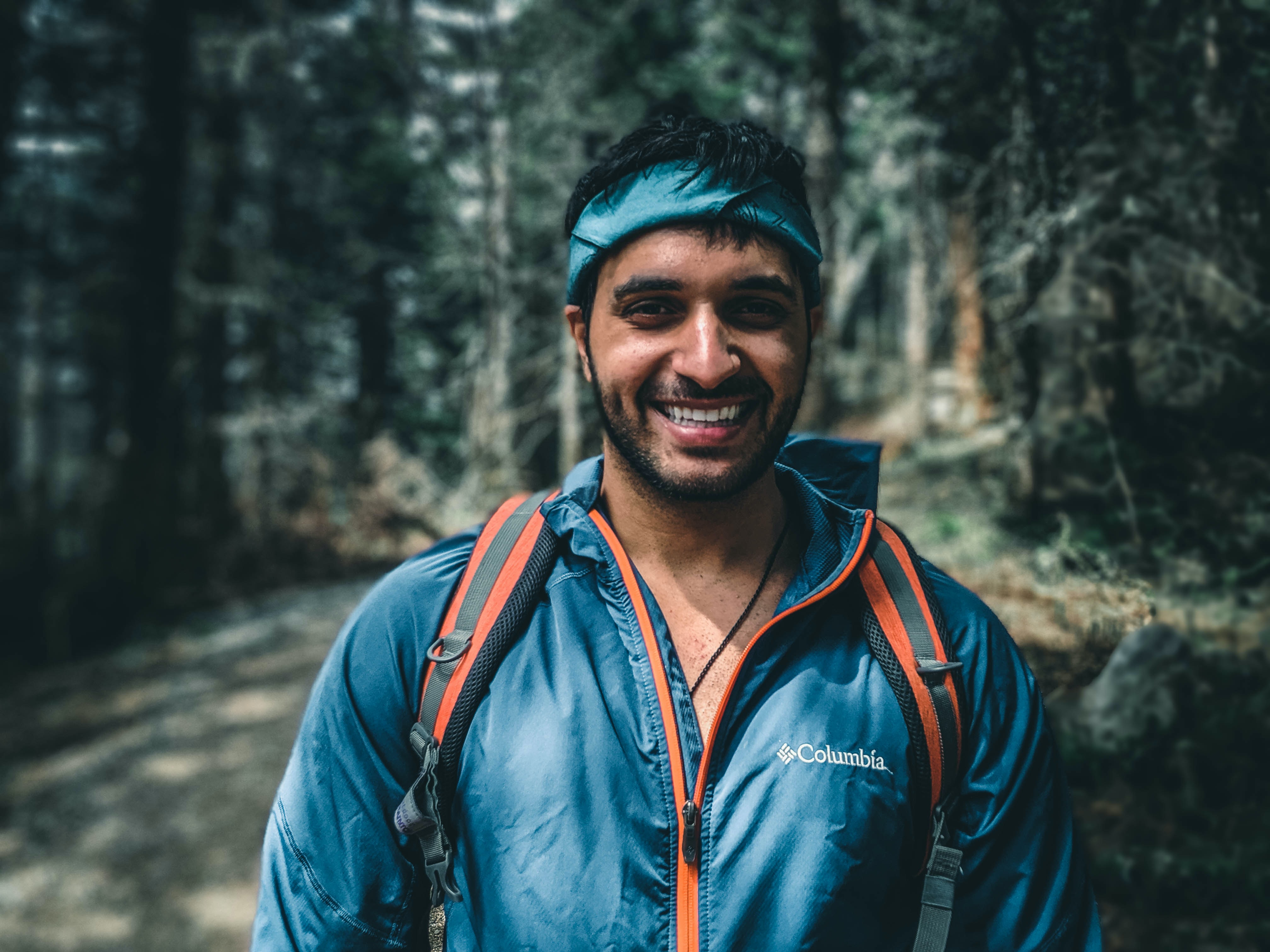 An attractive Indian man wearing a windbreaker and a bandana. He is smiling and on a wooded path hiking.