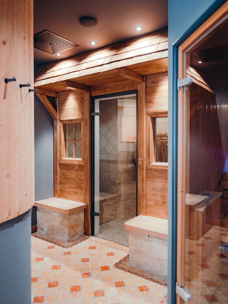 Eingang zu einer hölzernen Sauna mit Fenstern, Dachvorsprung und zweifarbigen Fliesen.