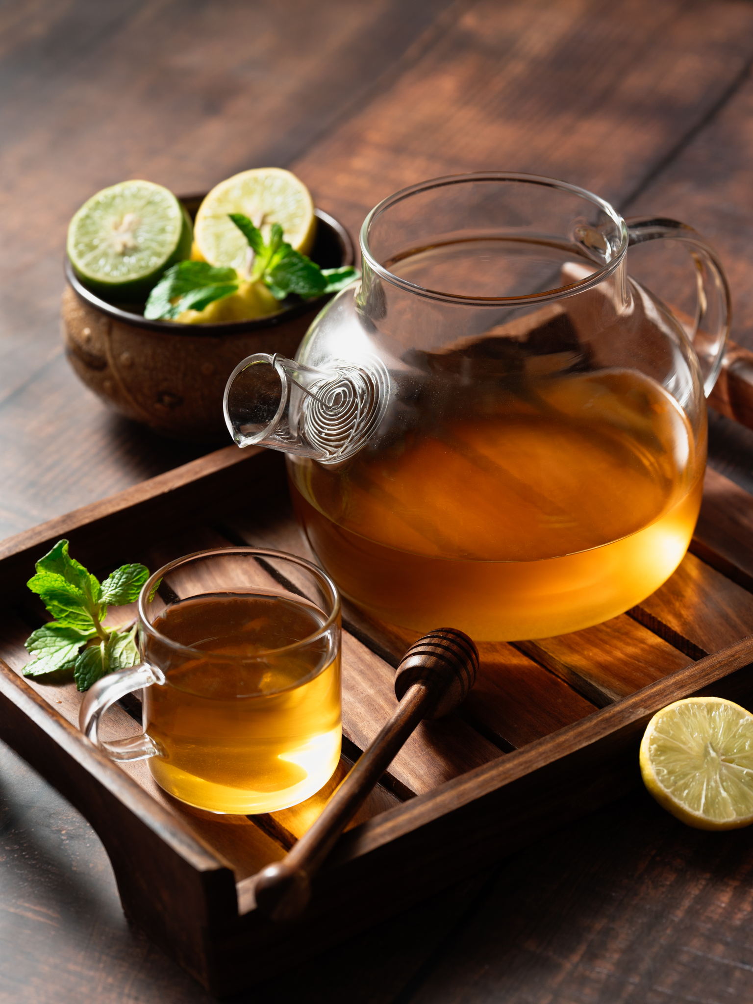 Herbal tea in a glass teapot and cup with lemon slices, mint leaves, and honey dipper on a wooden tray