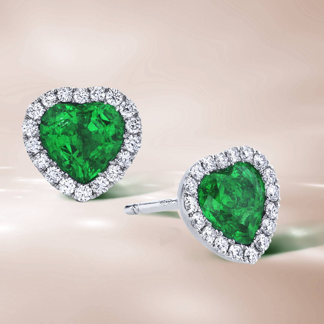 Heart shaped emerald stud earrings with diamond halos on a brown background.